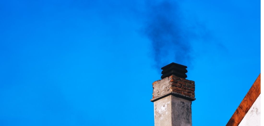 Fumée noire s’échappant d’une cheminée en briques sur le toit d’une maison sous un ciel bleu.